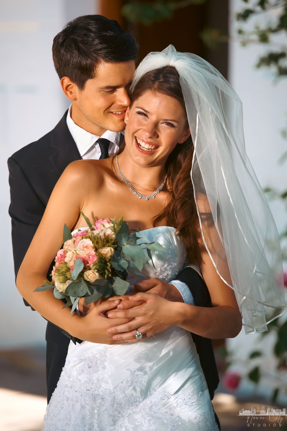 Bride in a veil smiling as the groom embraces her from behind